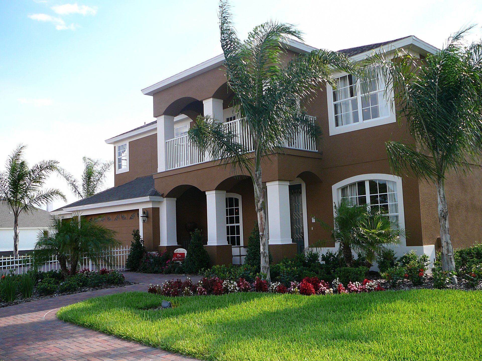 Two-story brown stucco house with white trim, a balcony, and palm trees in front yard.