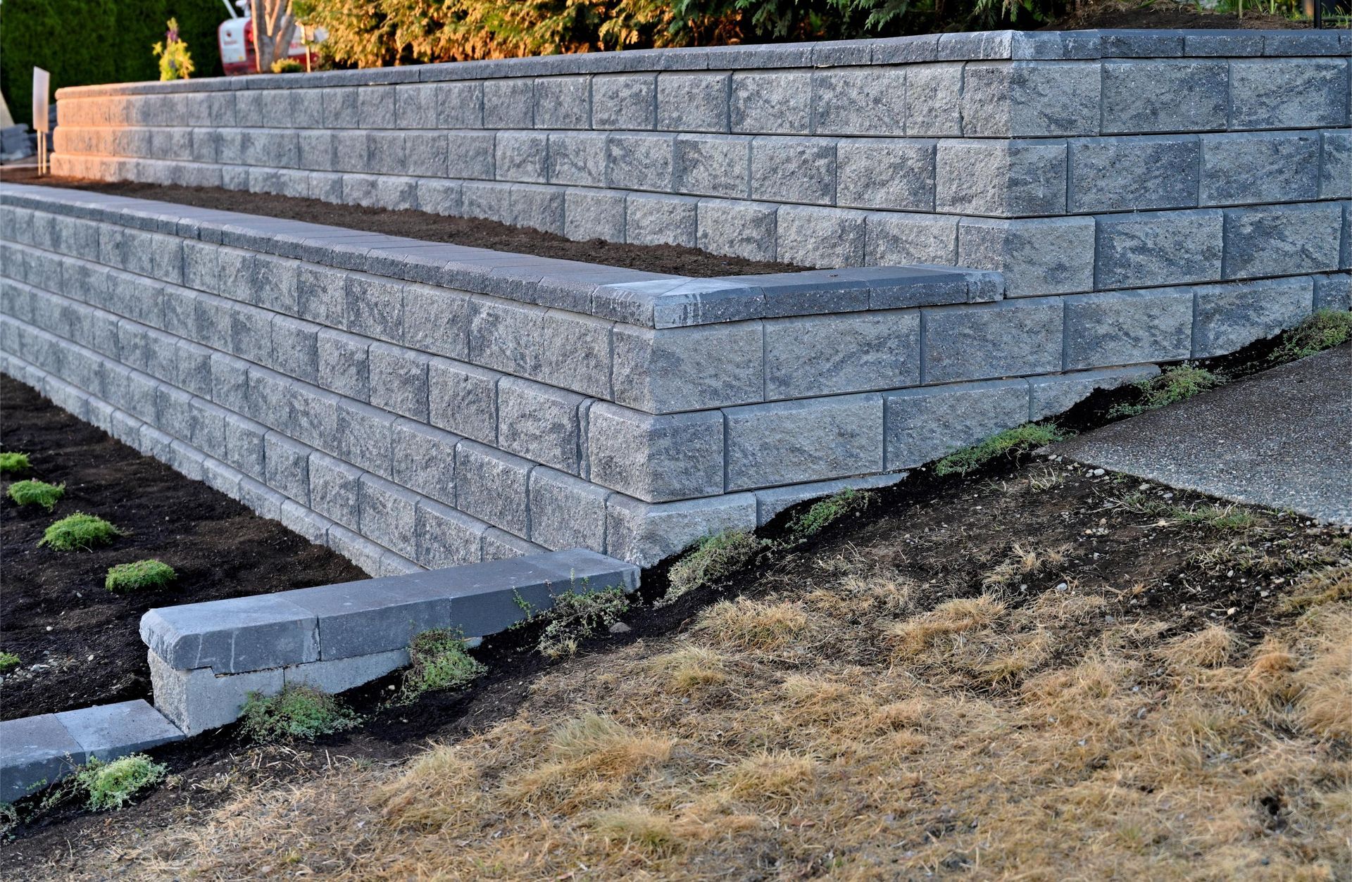 A multi-tiered retaining wall made of stacked grey concrete blocks bordering garden beds and a lawn.
