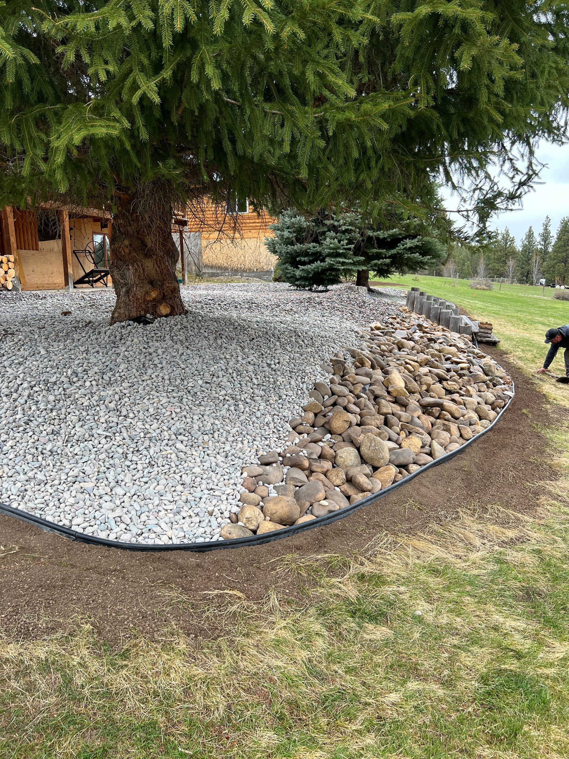Landscape bed with gray gravel and rounded rocks surrounding a tree. Bordered by brown mulch and lawn.