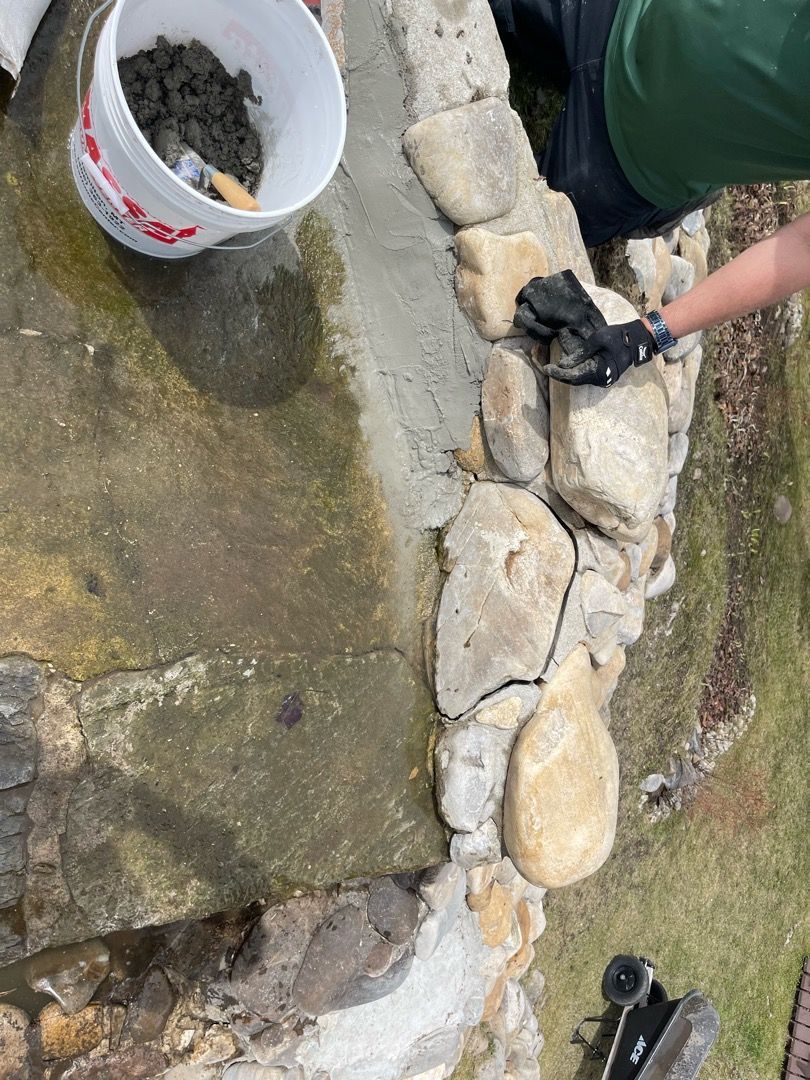 Person using a trowel to apply mortar between stones on a wall; a bucket of mortar and water are nearby.