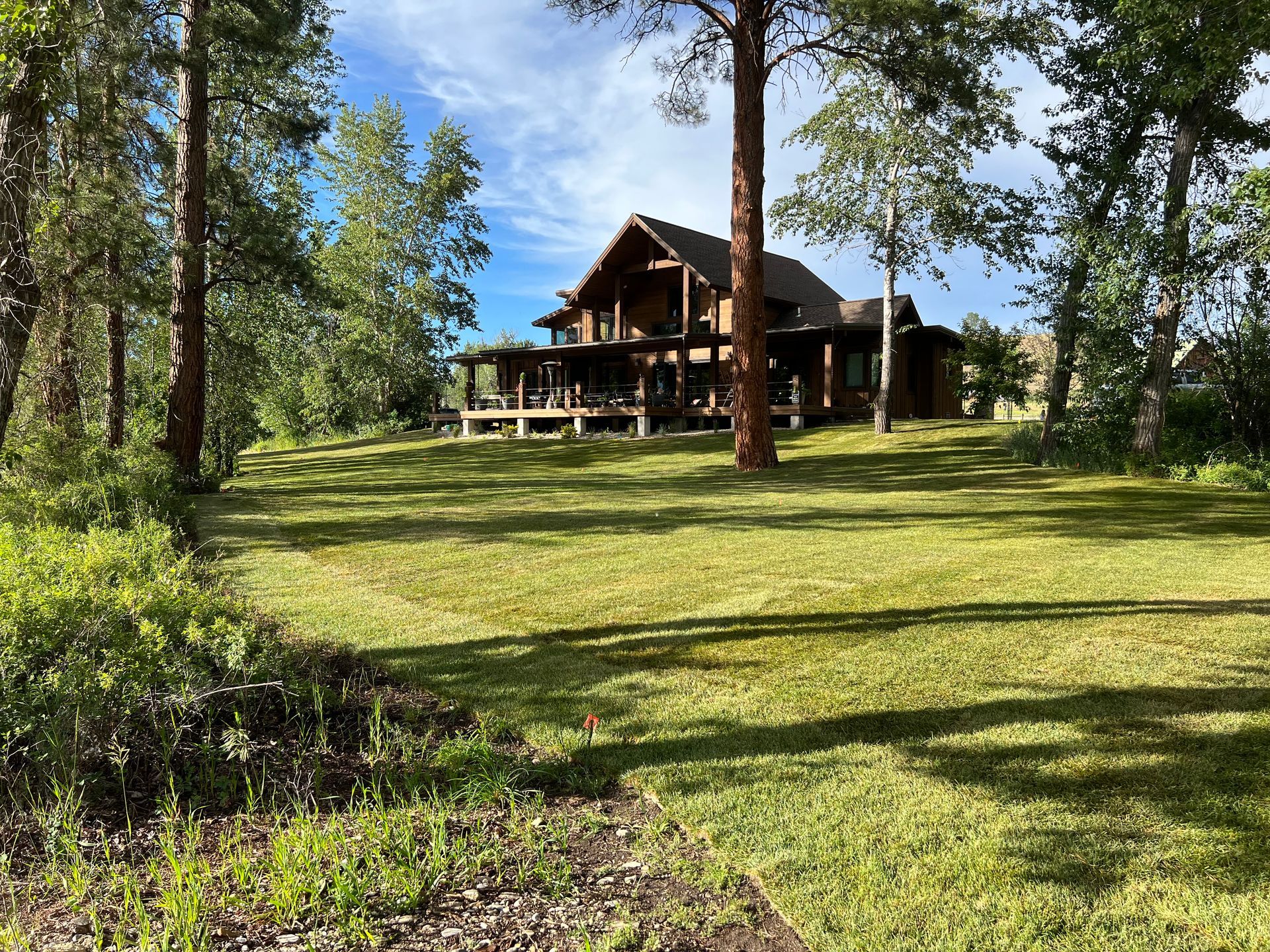 Large brown house with wrap-around porch, nestled among trees on a grassy lawn under a partly cloudy sky.