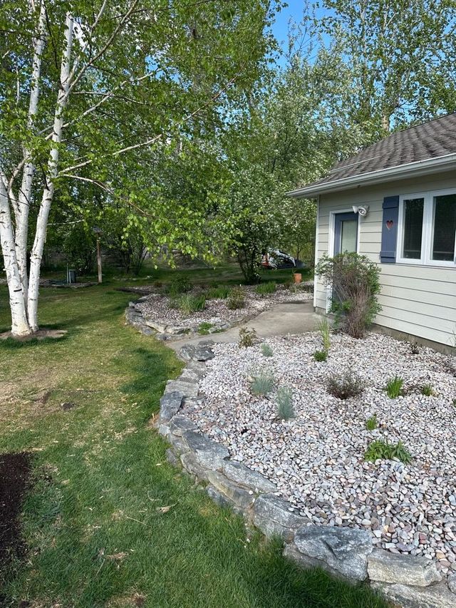 A house with a stone-edged flower bed filled with plants and gravel, beside a green lawn and trees.