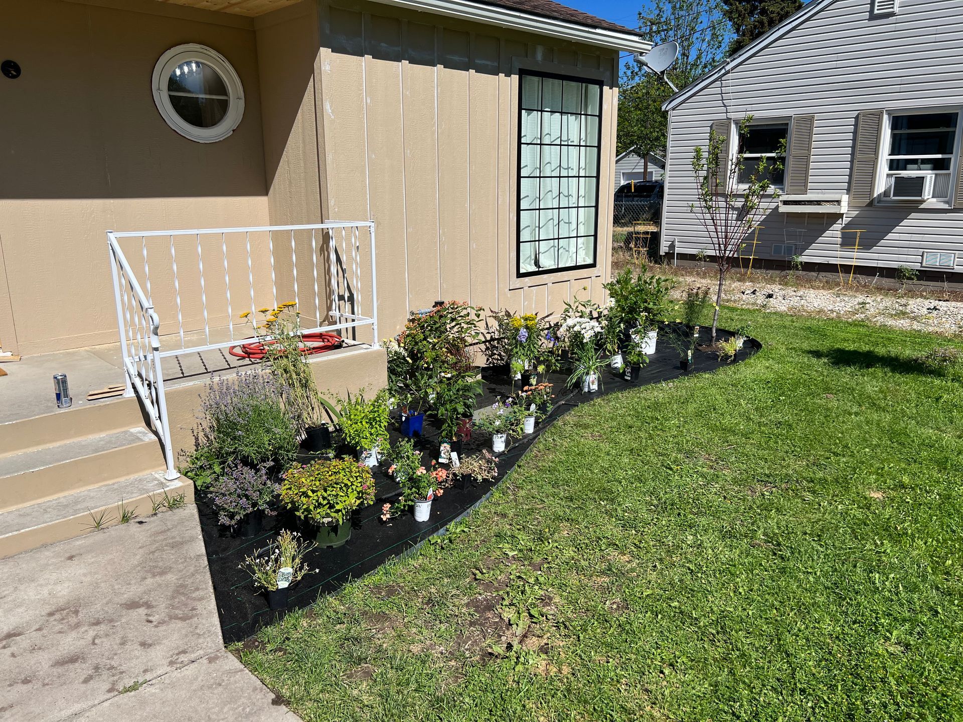 House exterior with garden bed filled with plants, mulch, and containers; sunny day.
