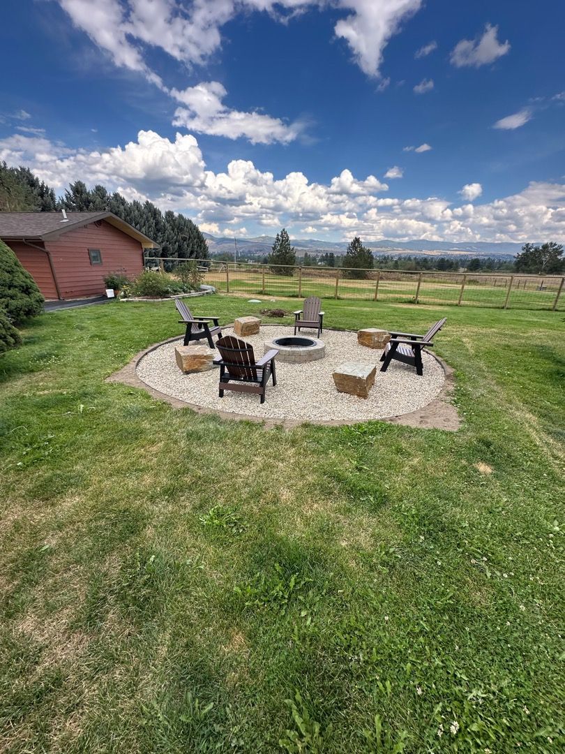 Fire pit area with chairs on a gravel base in a grassy yard, under a blue sky with clouds.
