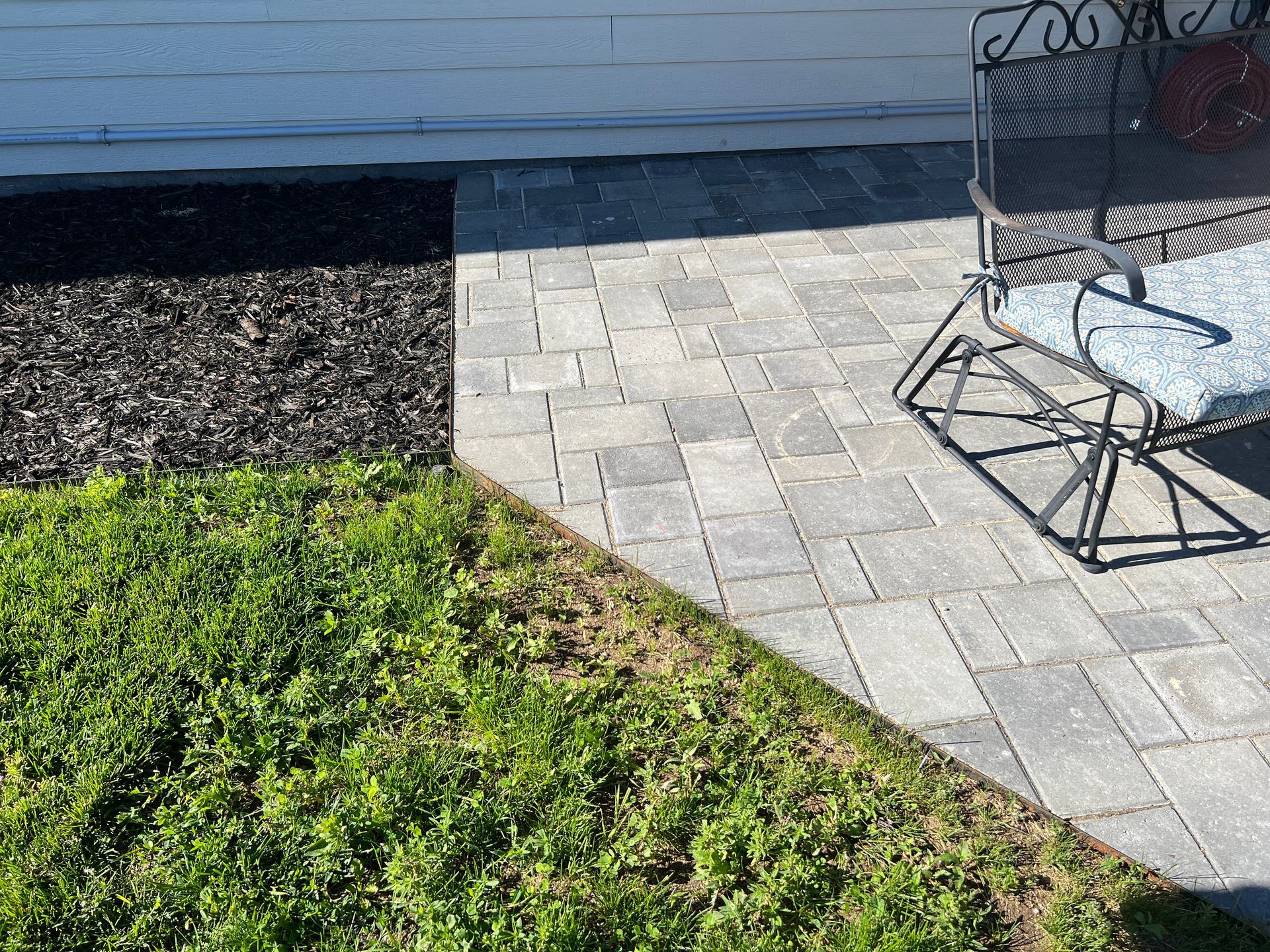 Patio pavers next to a grassy lawn and mulch. A wrought-iron bench sits on the pavers.
