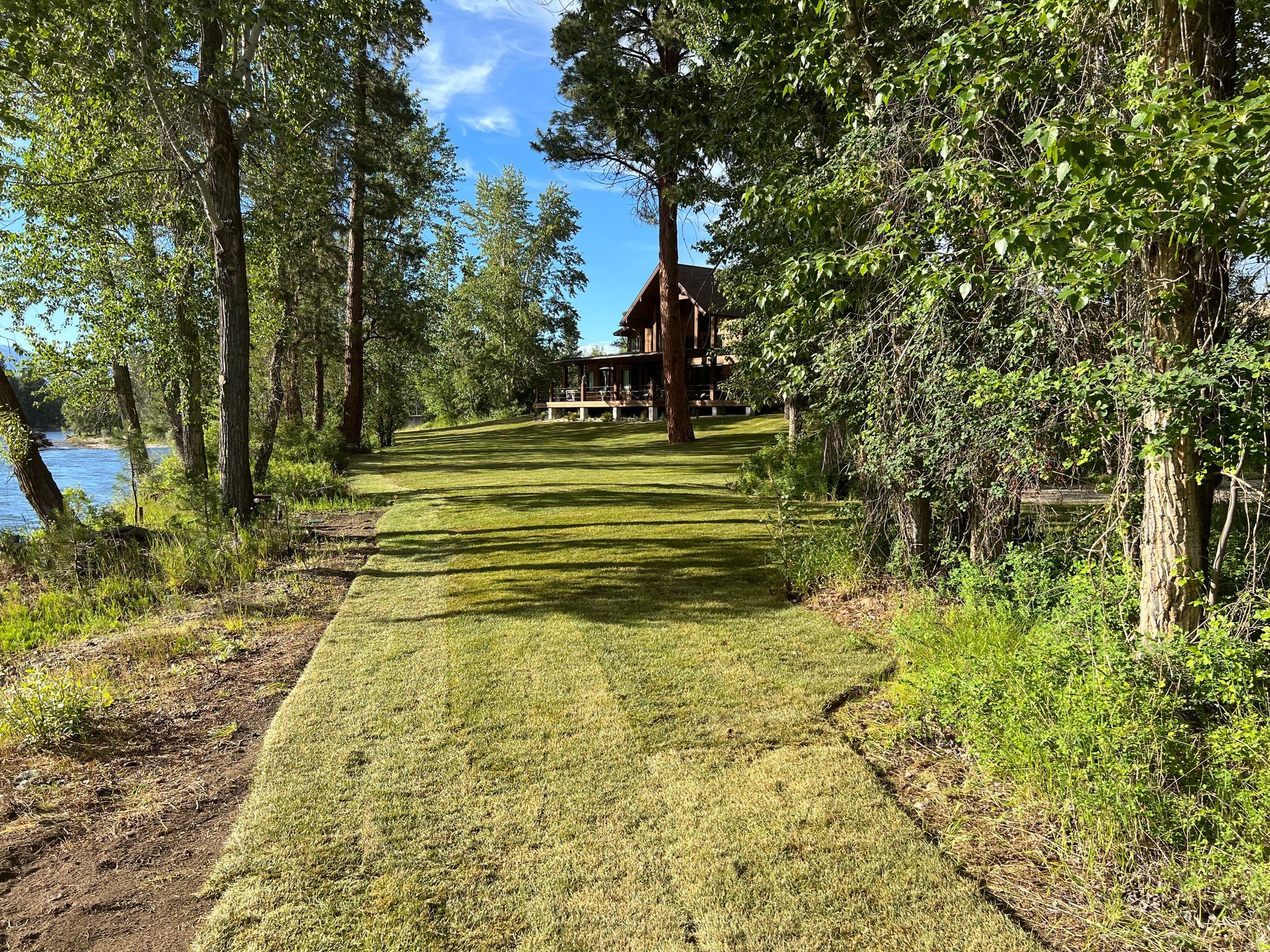 A grassy path leads to a house near a lake, surrounded by trees under a blue sky.