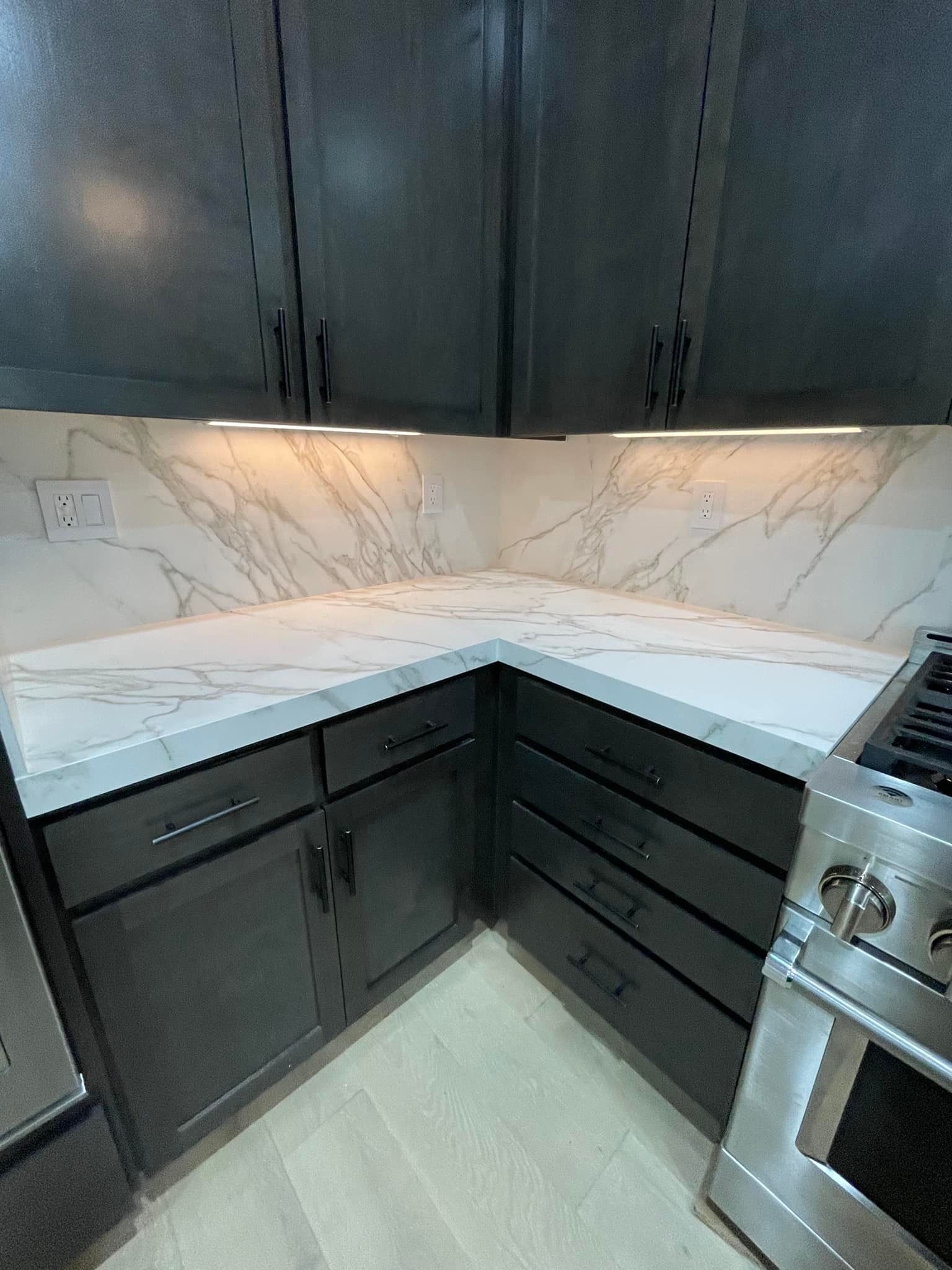 Dark gray kitchen cabinets and white countertops with a marble pattern. Built-in oven, bright lights under cabinets.