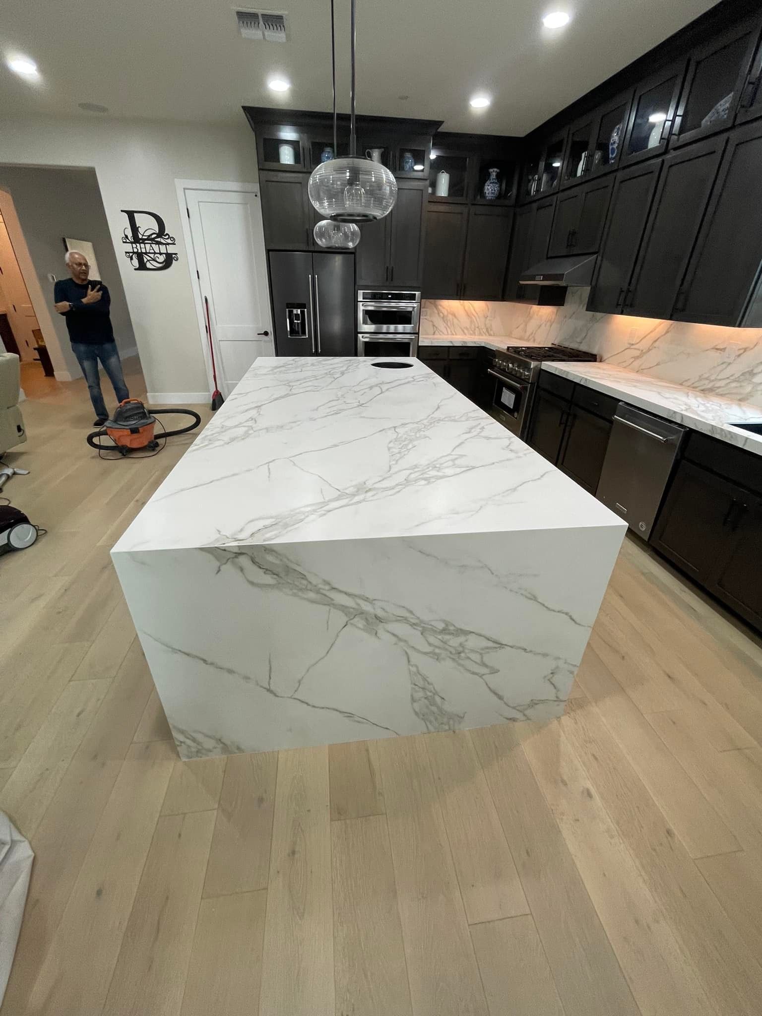 Modern kitchen with white marble island, dark cabinets, stainless steel appliances, and pendant lights.