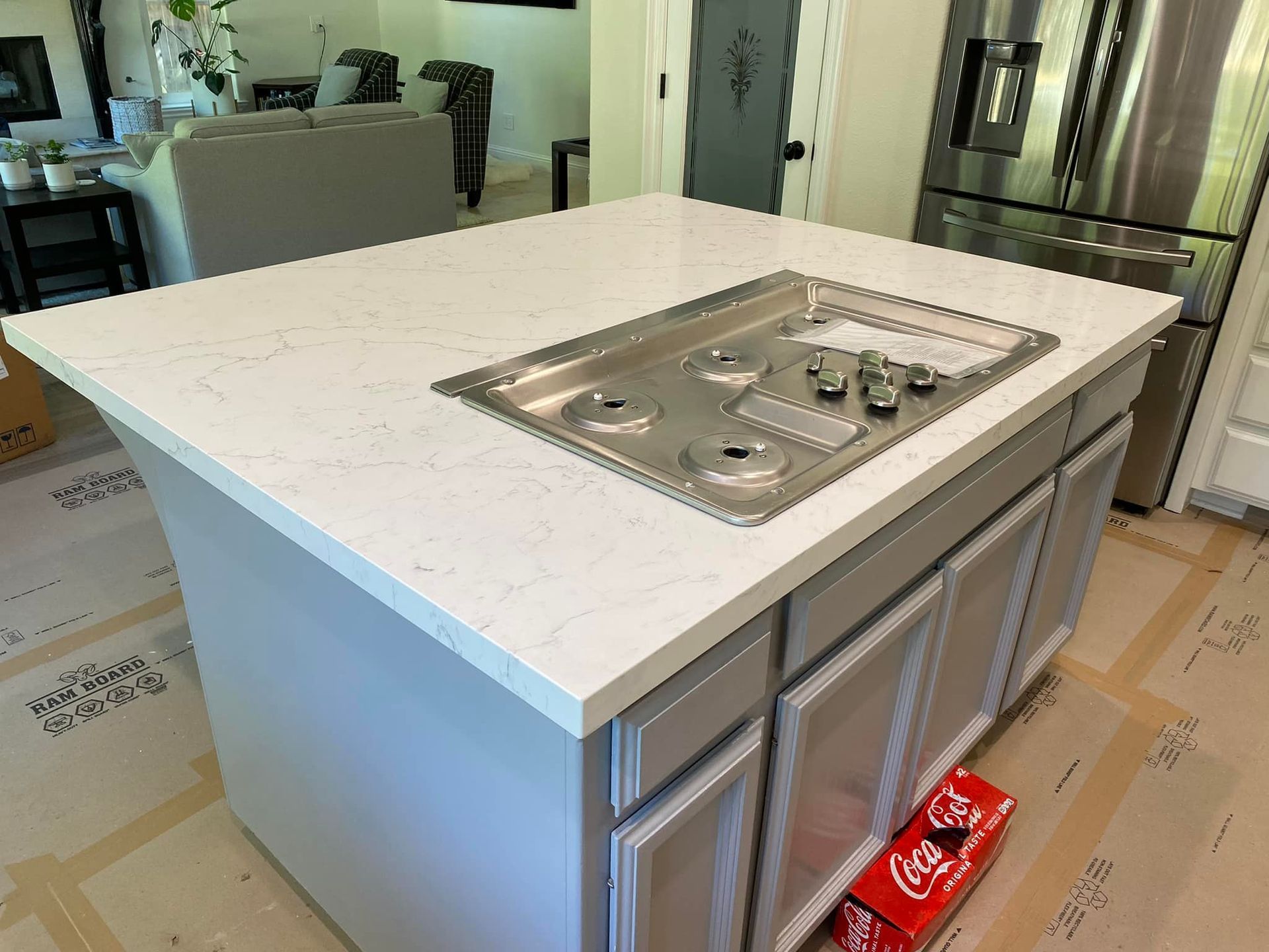 Kitchen island with cooktop, grey cabinets, and white countertop. A soda box sits on the floor.