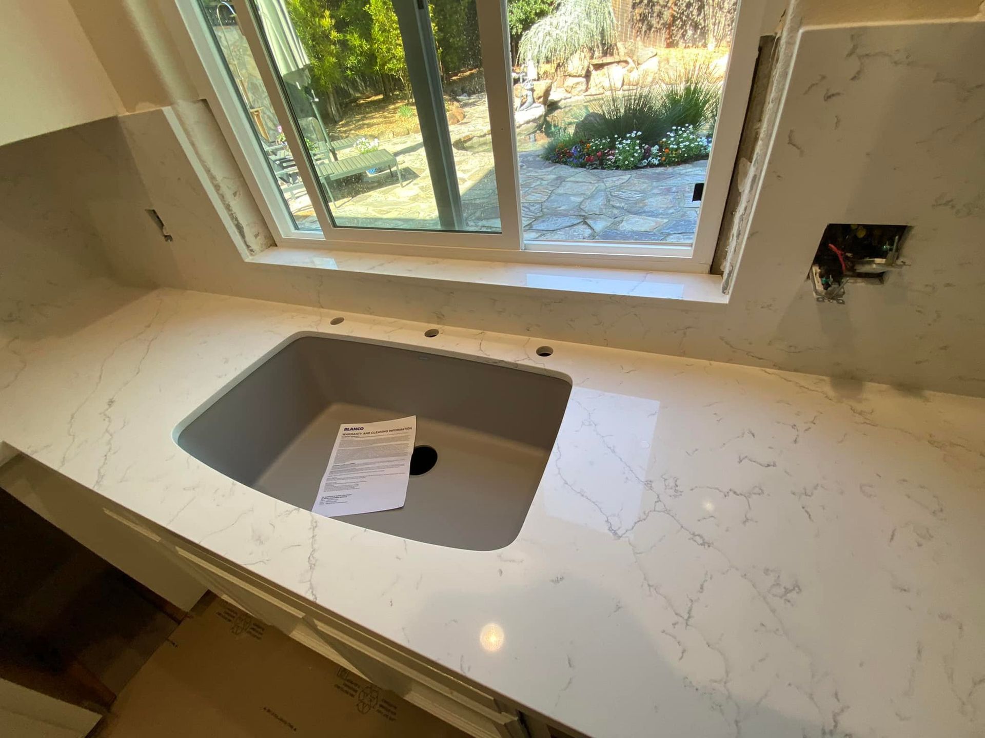 White countertop with a sink, near a window overlooking a garden.