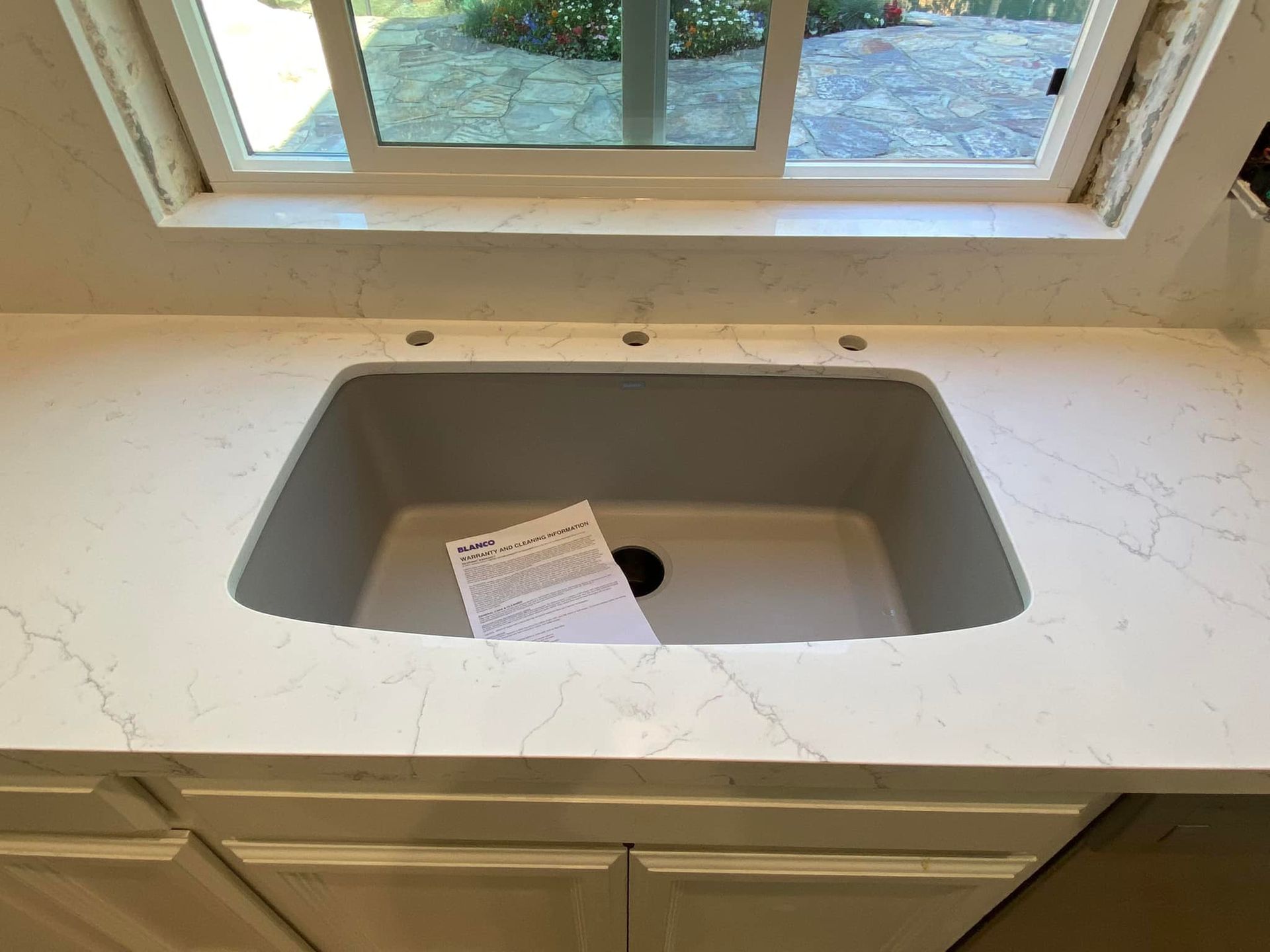 A newly installed kitchen sink with a white countertop, above a white cabinet and below a window.