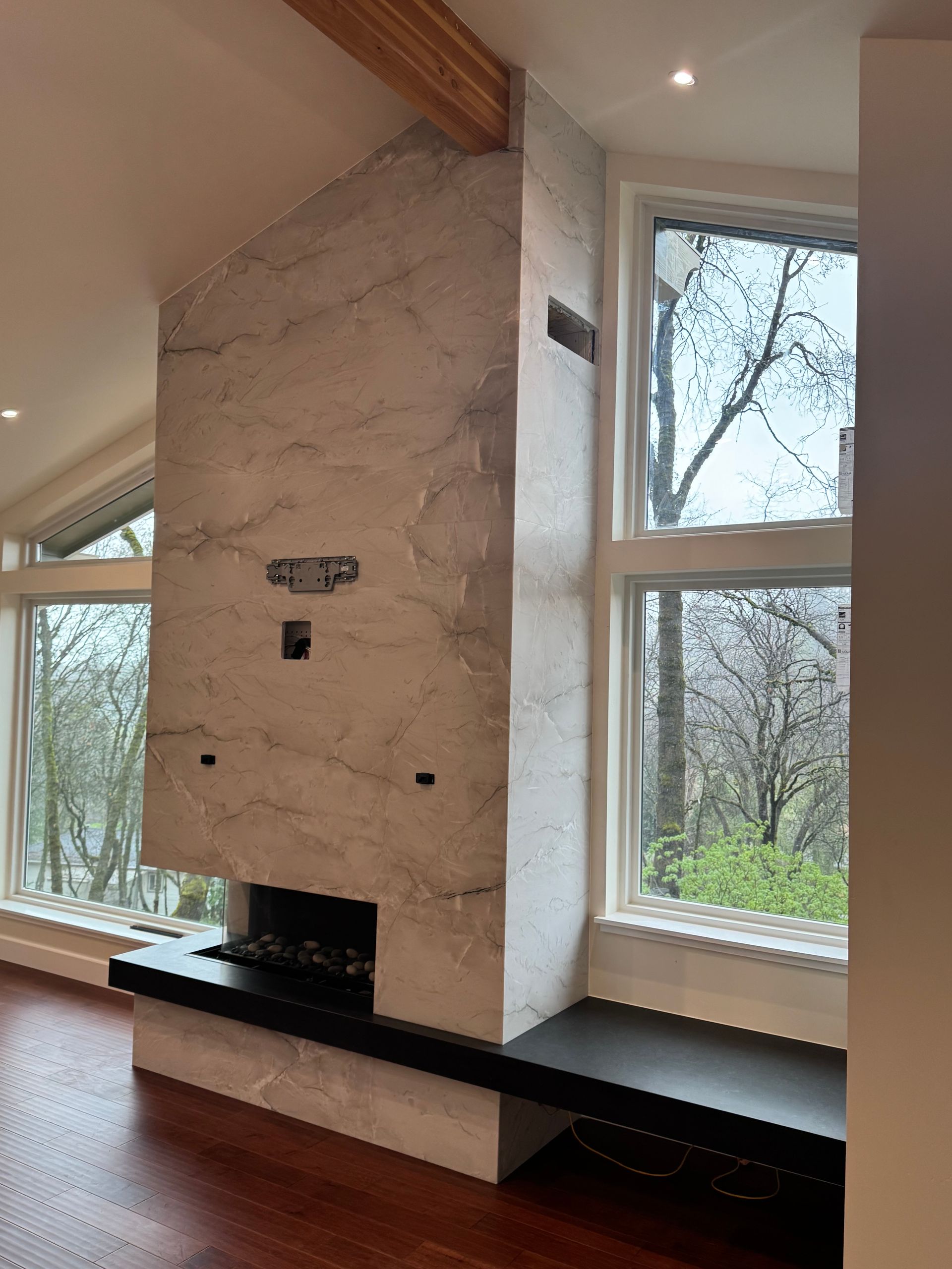 Modern fireplace with marble-like finish and black hearth. Large windows, wood floors, and high ceiling.
