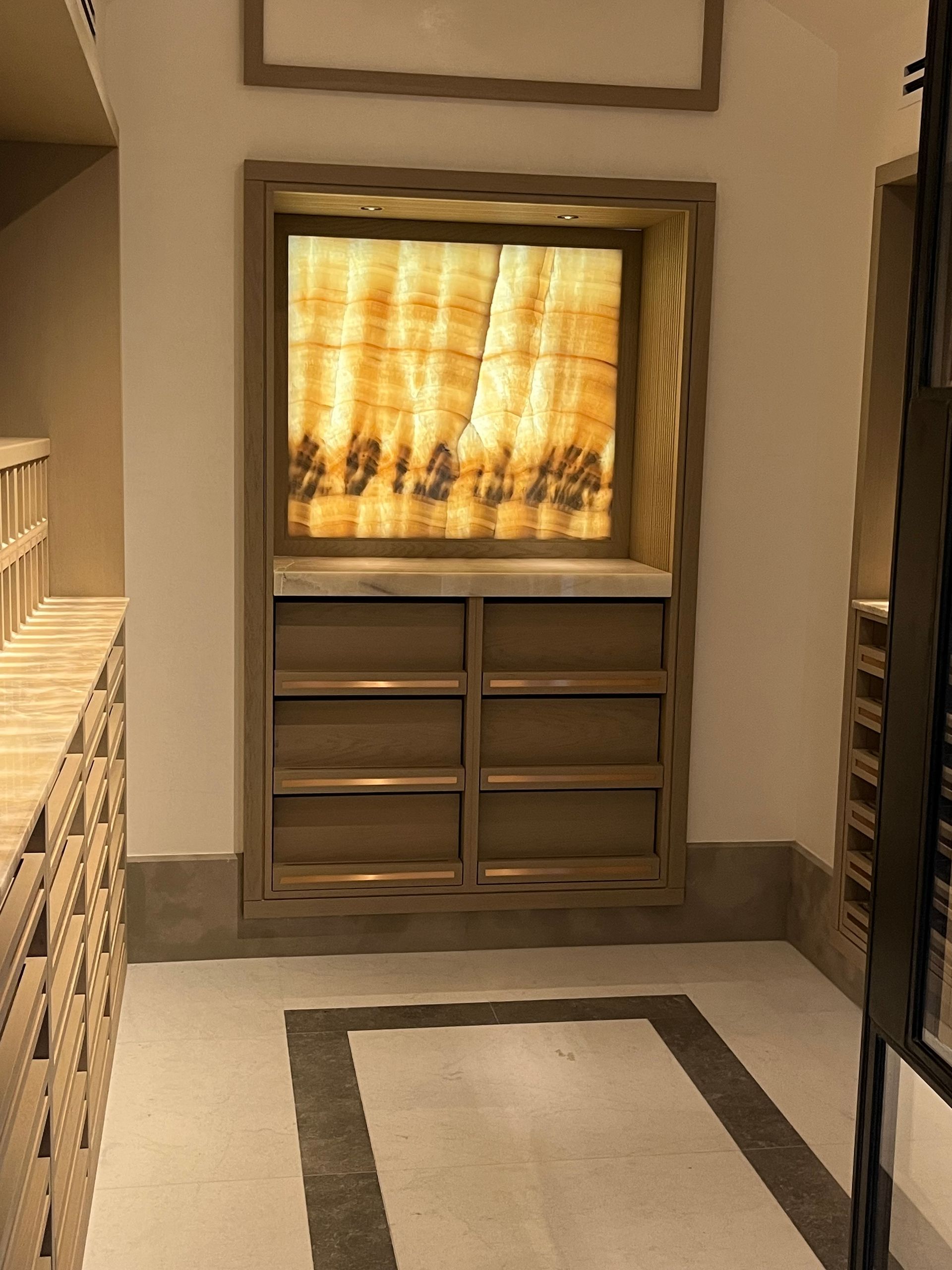 A wall niche with a lit, layered amber panel above six drawers in a hallway.