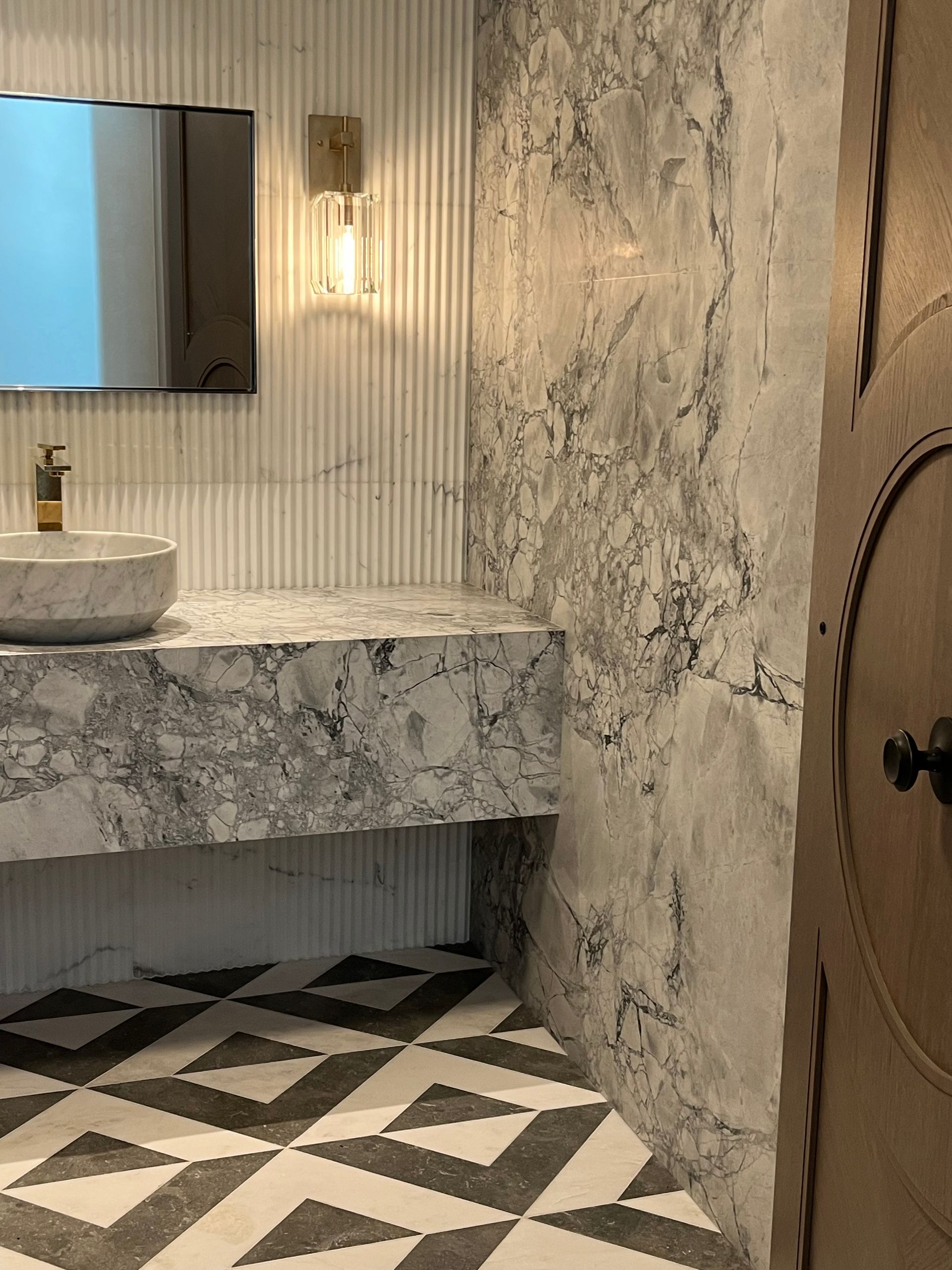 Bathroom with marble sink and countertop, textured gray walls, geometric tile floor, brass sconce, and a wooden door.