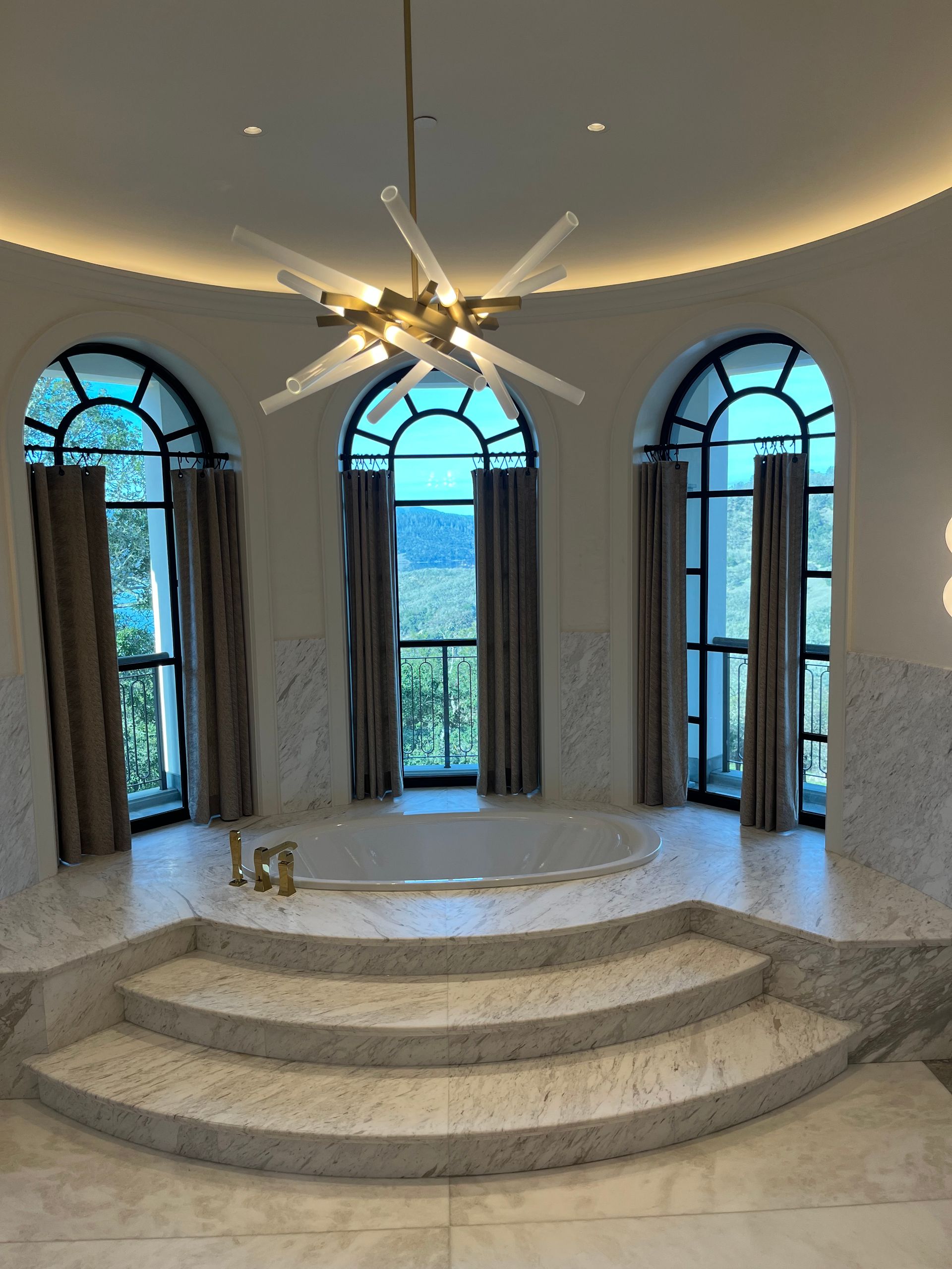 Bathroom with three arched windows, a round tub, and steps. A starburst chandelier hangs from the ceiling.