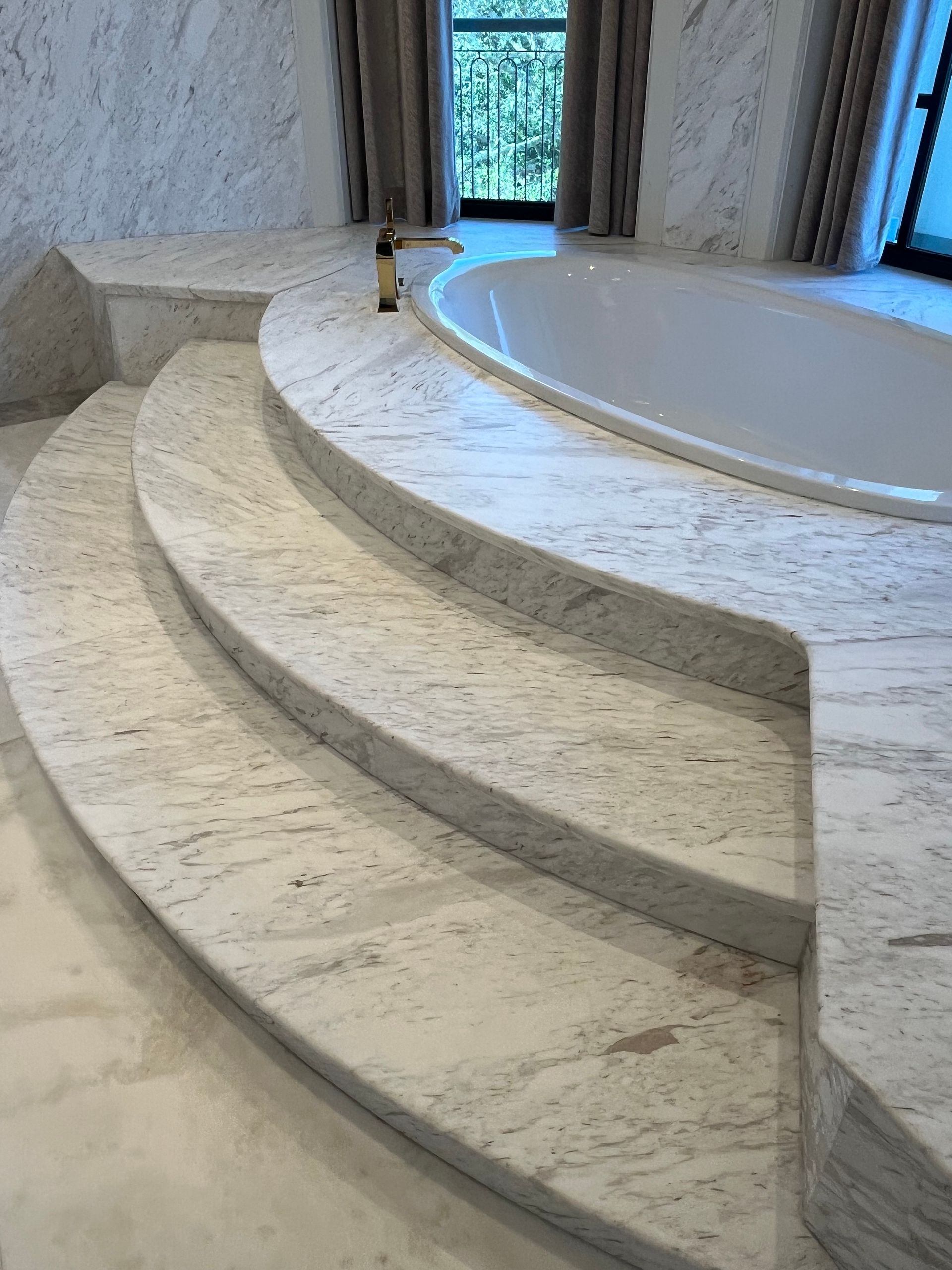 Marble bathroom with steps leading to a built-in oval bathtub. Gold faucet, window with curtains.