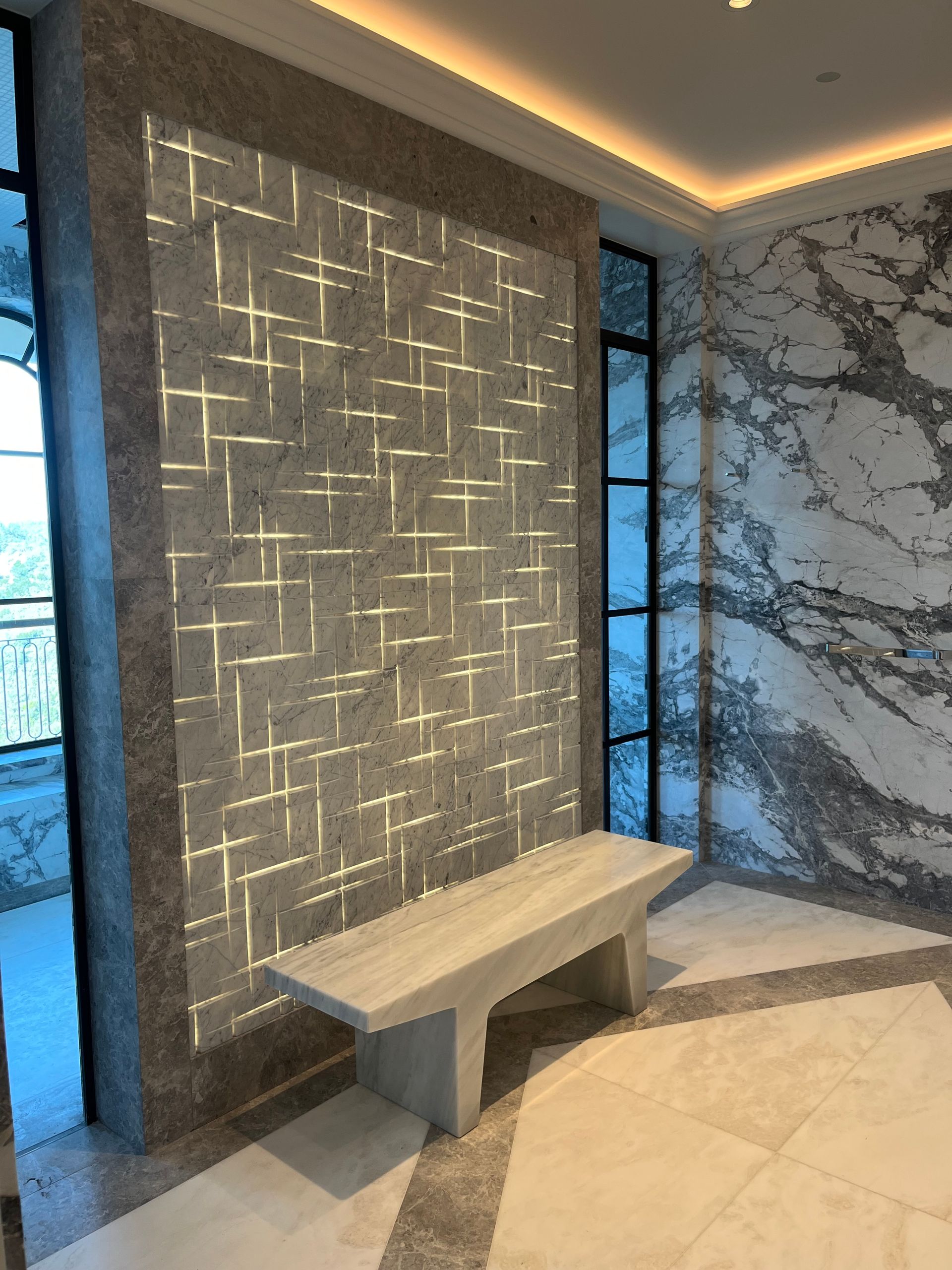 A modern entryway features a bench beneath a gray wall with a silver geometric design and marble walls.