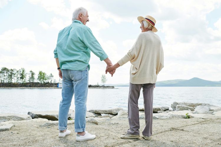 Two people holding hands on a rocky shoreline beside calm water under a bright sky
