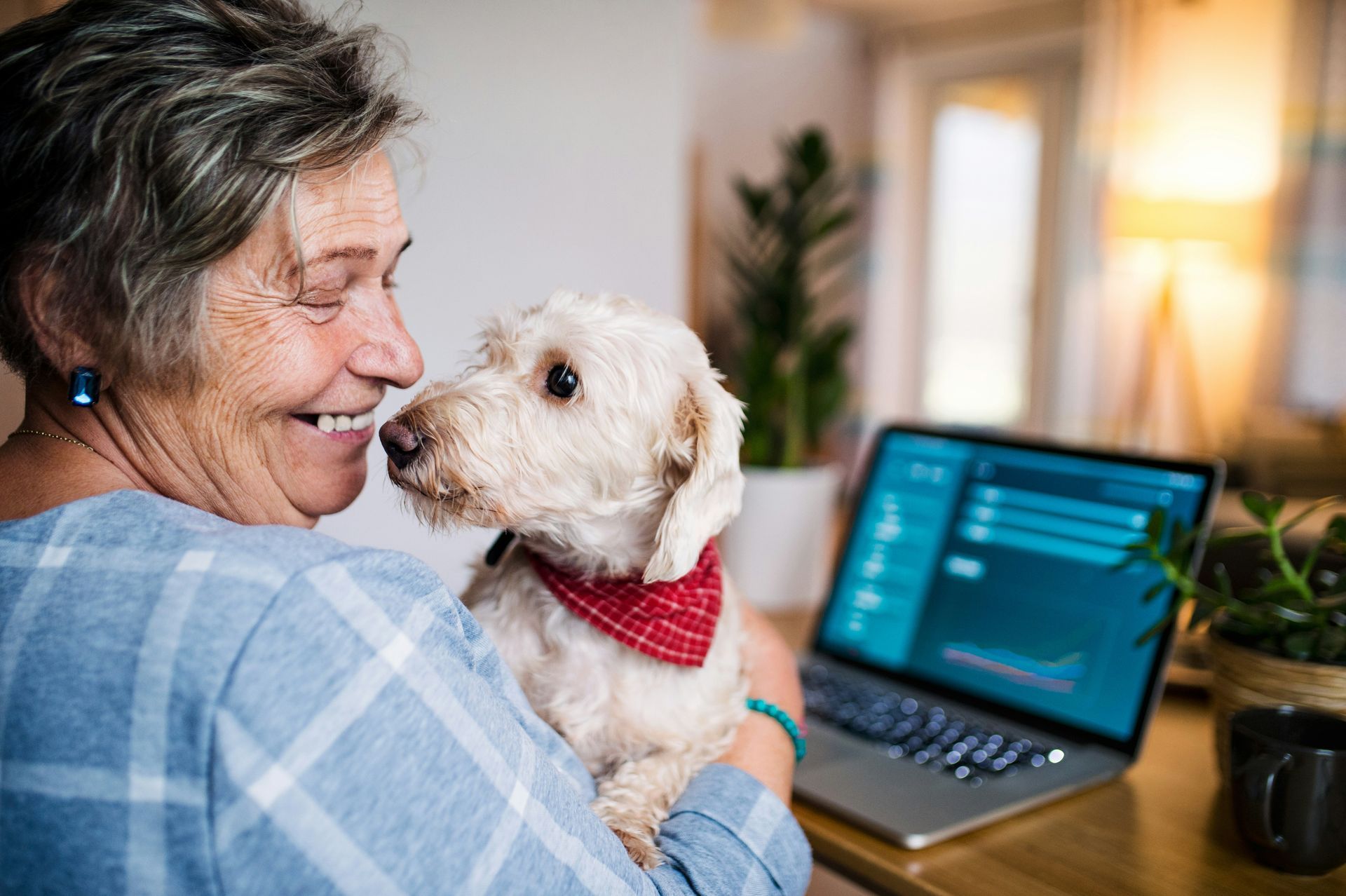 Woman cuddles a small white dog with a red bandana beside a laptop in a cozy home setting