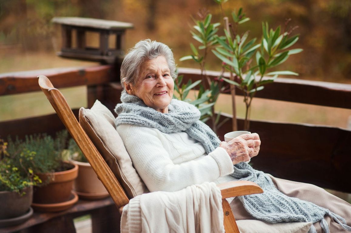 Older woman relaxing on a wooden chair on a porch, holding a plant and smiling amid potted greenery.