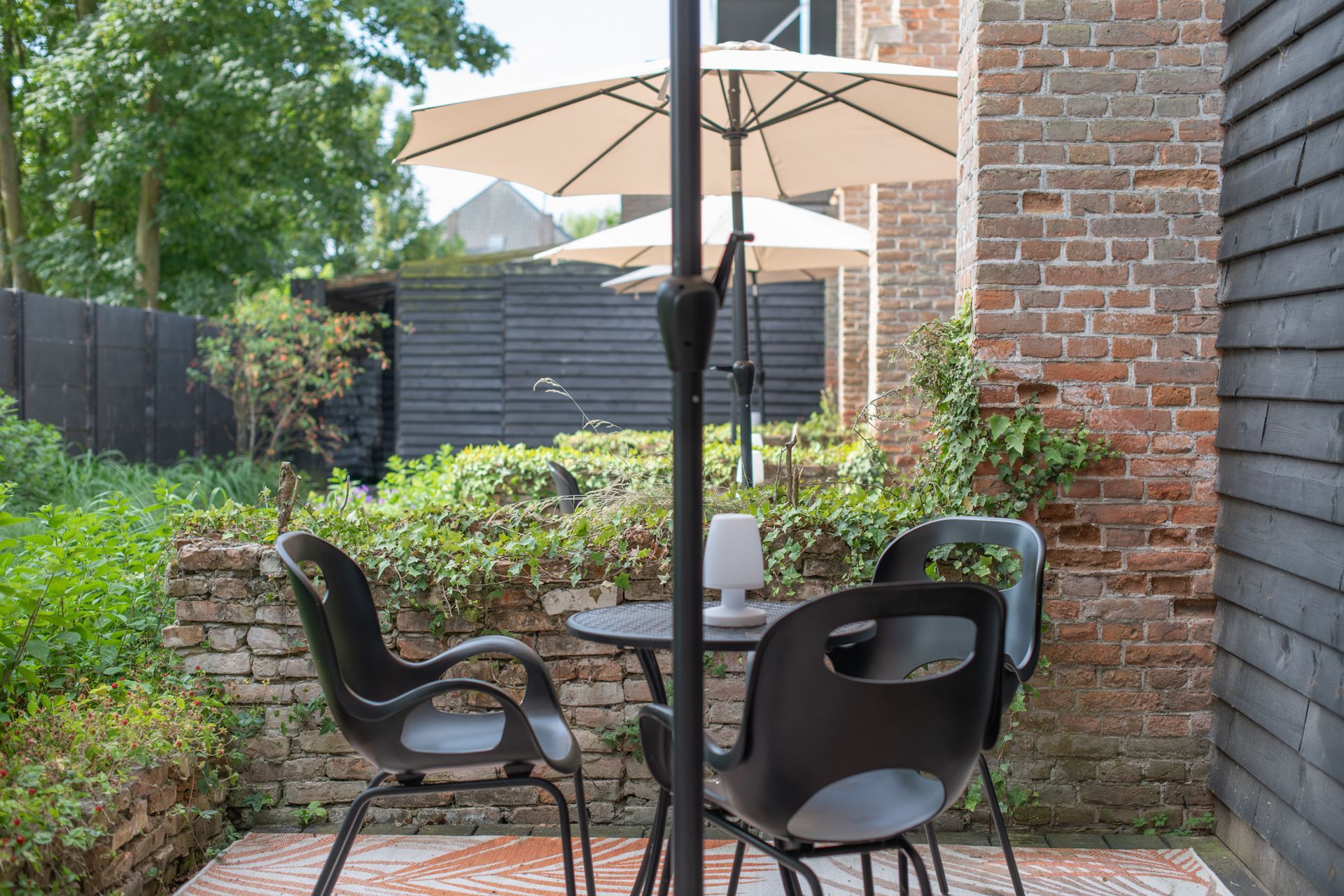 Een kleine terrastafel met vier zwarte stoelen op een vloerkleed met patroon, in de schaduw van een parasol naast een bakstenen muur en tuin.