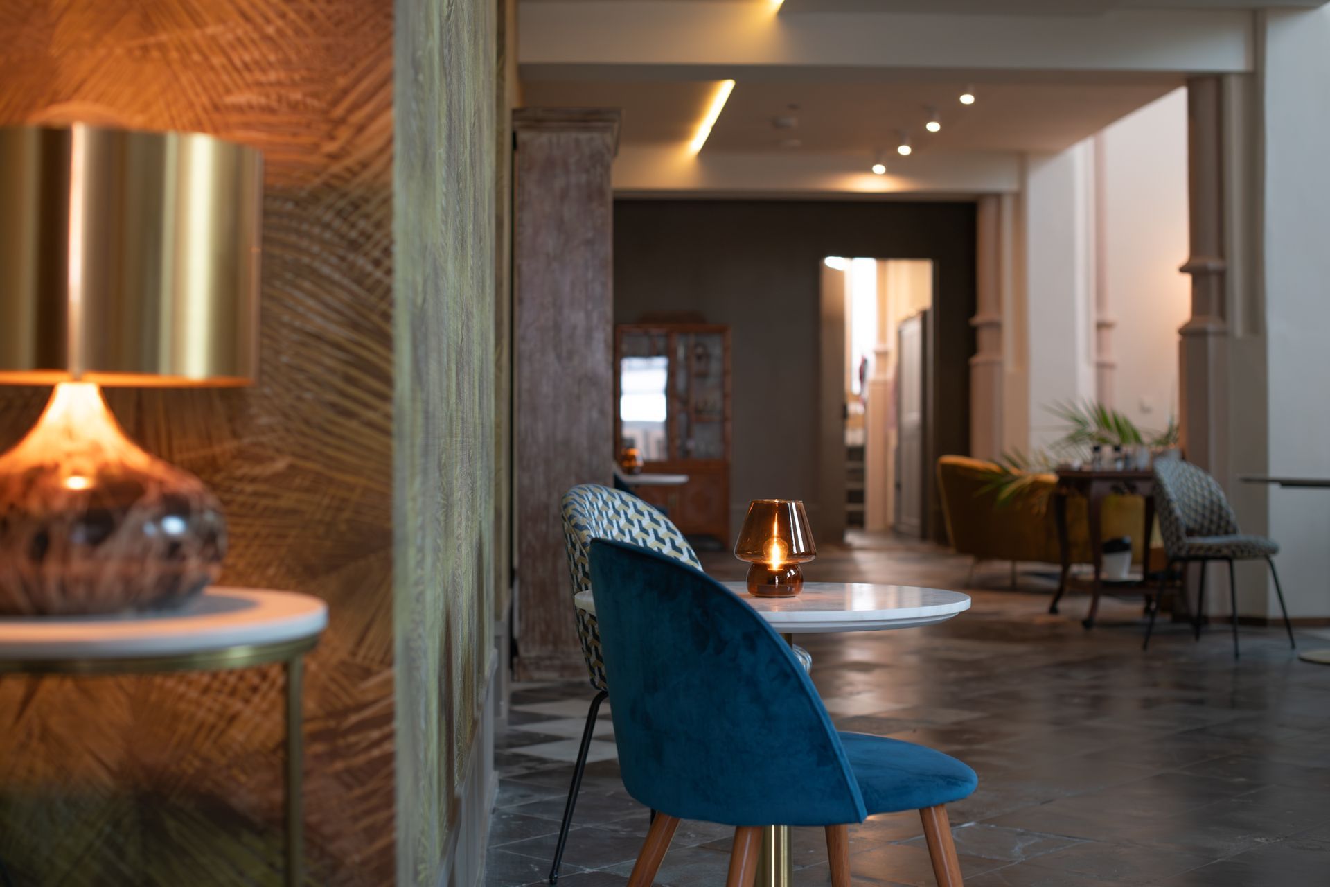 Een sfeervolle, ietwat gedempte hotellobby met een gouden lamp op de voorgrond, een blauwe fluwelen stoel en zachte, sfeervolle verlichting.
