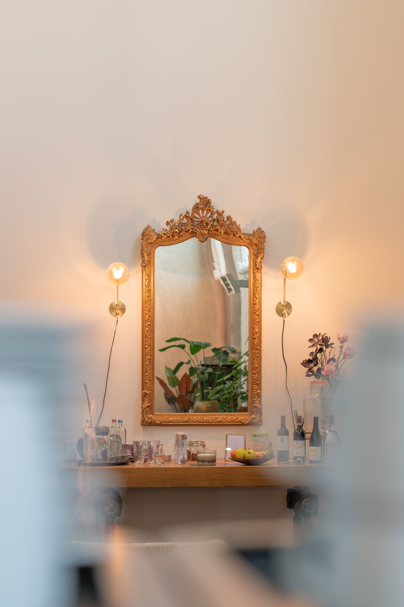 Een sierlijke spiegel met gouden lijst hangt boven een houten plank met planten, glazen flessen en twee sfeervolle wandlampen.