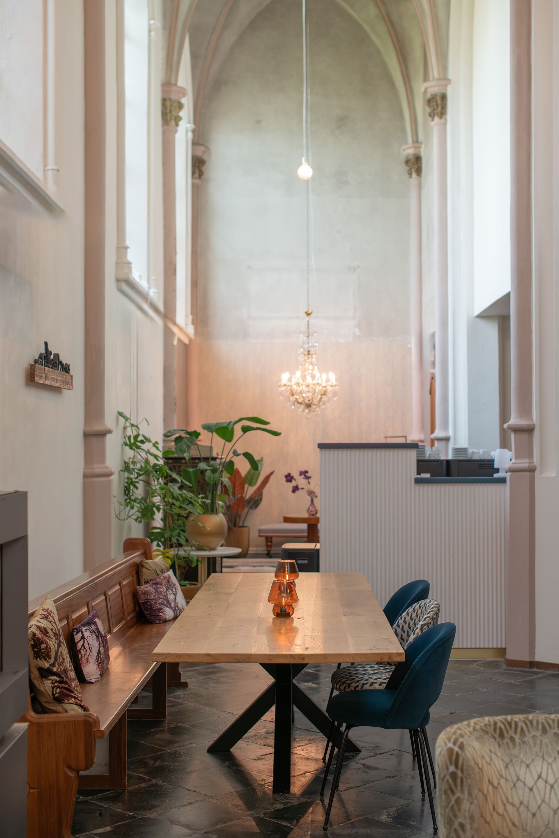 Een lange eettafel met een houten blad en een zwart onderstel, in een lichte kamer met hoge gewelfde plafonds en een kroonluchter.