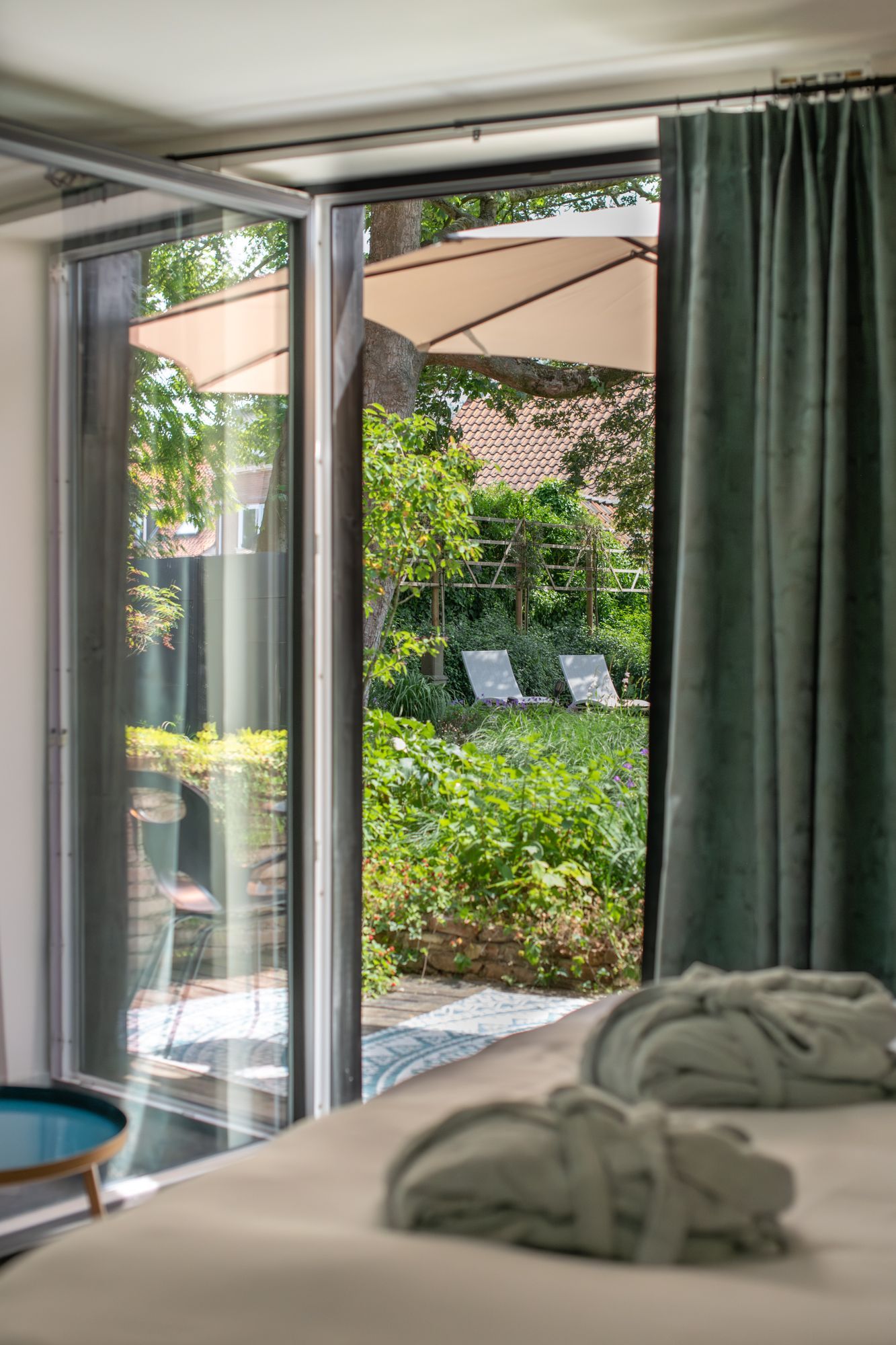 Een uitzicht vanuit een hotelbed, met twee opgevouwen badjassen, en door een glazen deur naar een zonnige, groene tuinpatio.