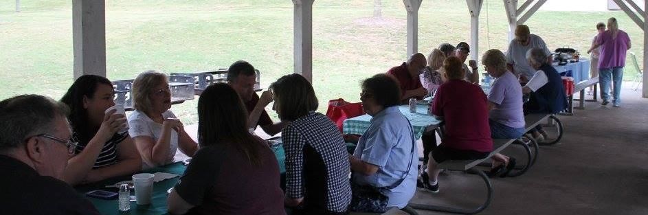 People seated at picnic tables under a pavilion, eating and talking in a shaded outdoor area.