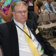Man in a black suit and yellow tie seated at an indoor event, looking toward the camera