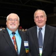 Two men in suits posing together at an event, one with a colorful tie and both wearing name badges.
