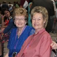 Two women smiling at a crowded indoor event, wearing blue and pink blouses.