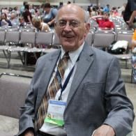 Smiling man in a gray suit with a lanyard seated in an auditorium with empty chairs behind him