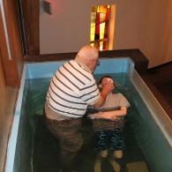 Man baptizing a person in a small indoor baptismal pool with stained glass in the background