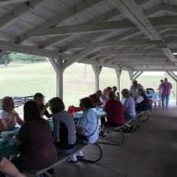 People seated at picnic tables under a wooden pavilion in a park