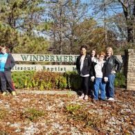 People standing by a Windermere Missouri Baptist sign in a wooded outdoor setting.