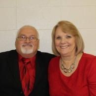 Two smiling people seated side by side, wearing red and black against a light wall.