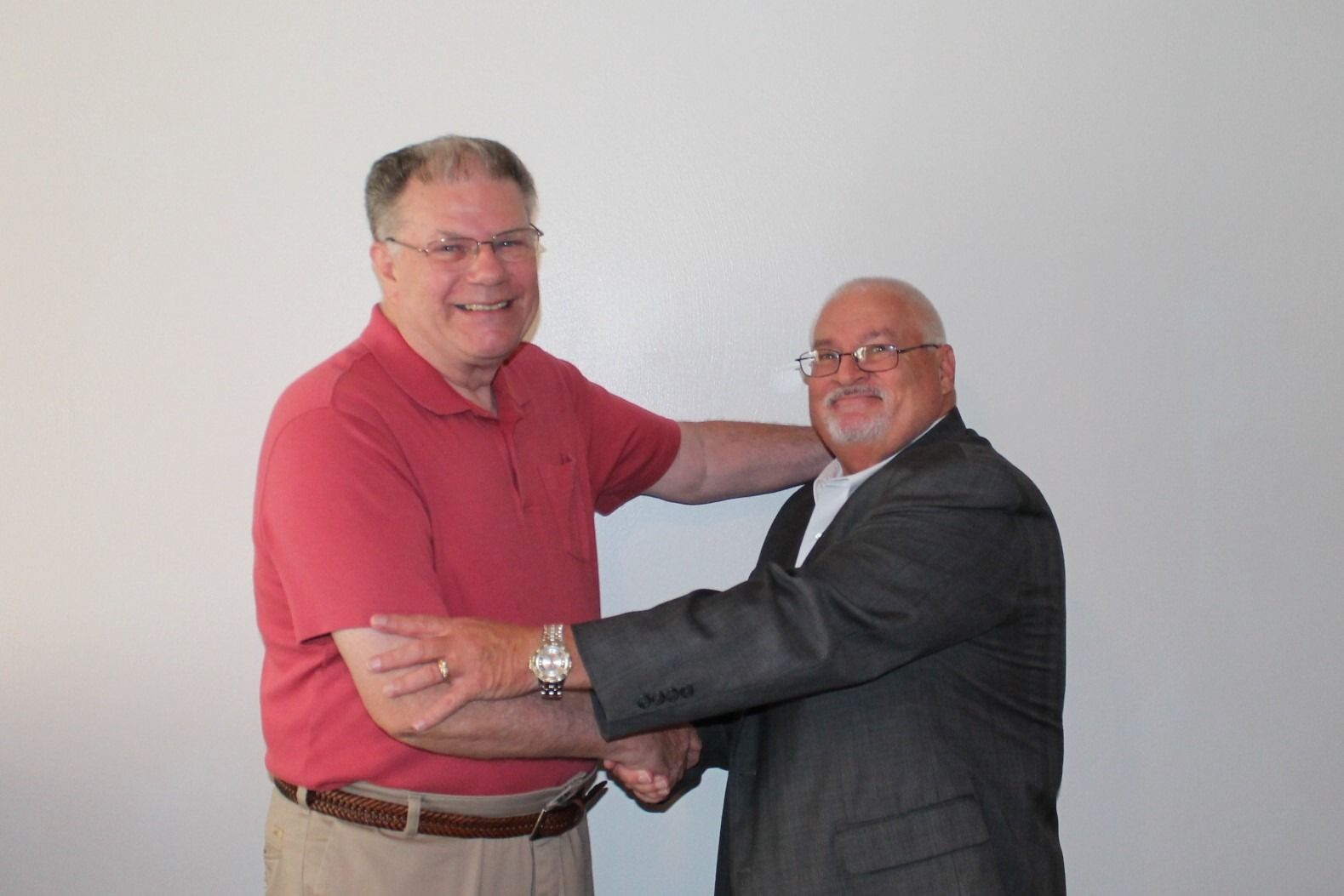 Two men smiling and embracing, one in a red shirt and one in a dark suit, against a plain wall.