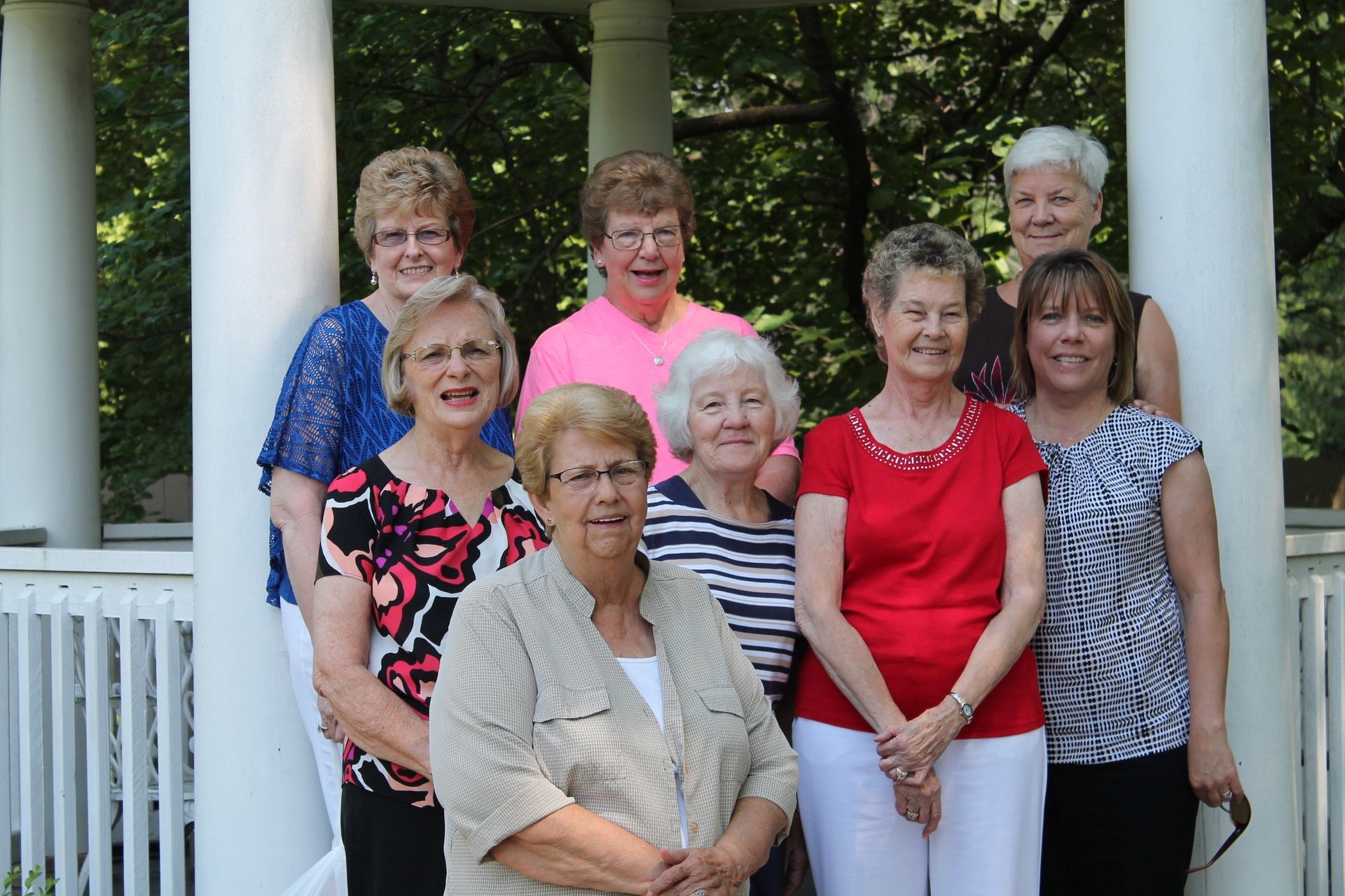 Eight people posing together outdoors on a porch with white columns and railings