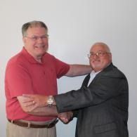 Two men smiling and shaking hands indoors, one in a red shirt and one in a dark suit.