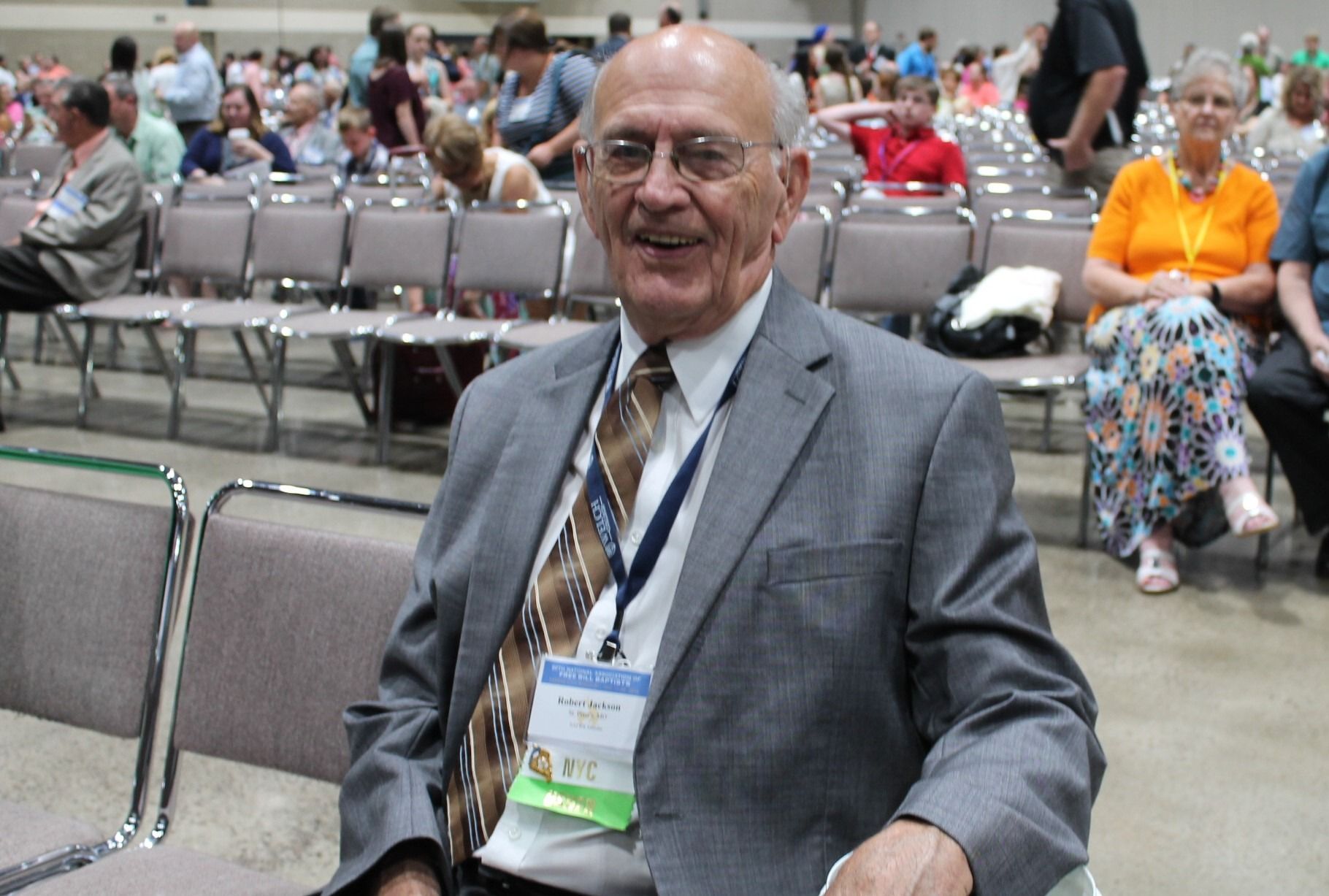 Smiling man in gray suit sitting in an auditorium, with rows of chairs and people in the background