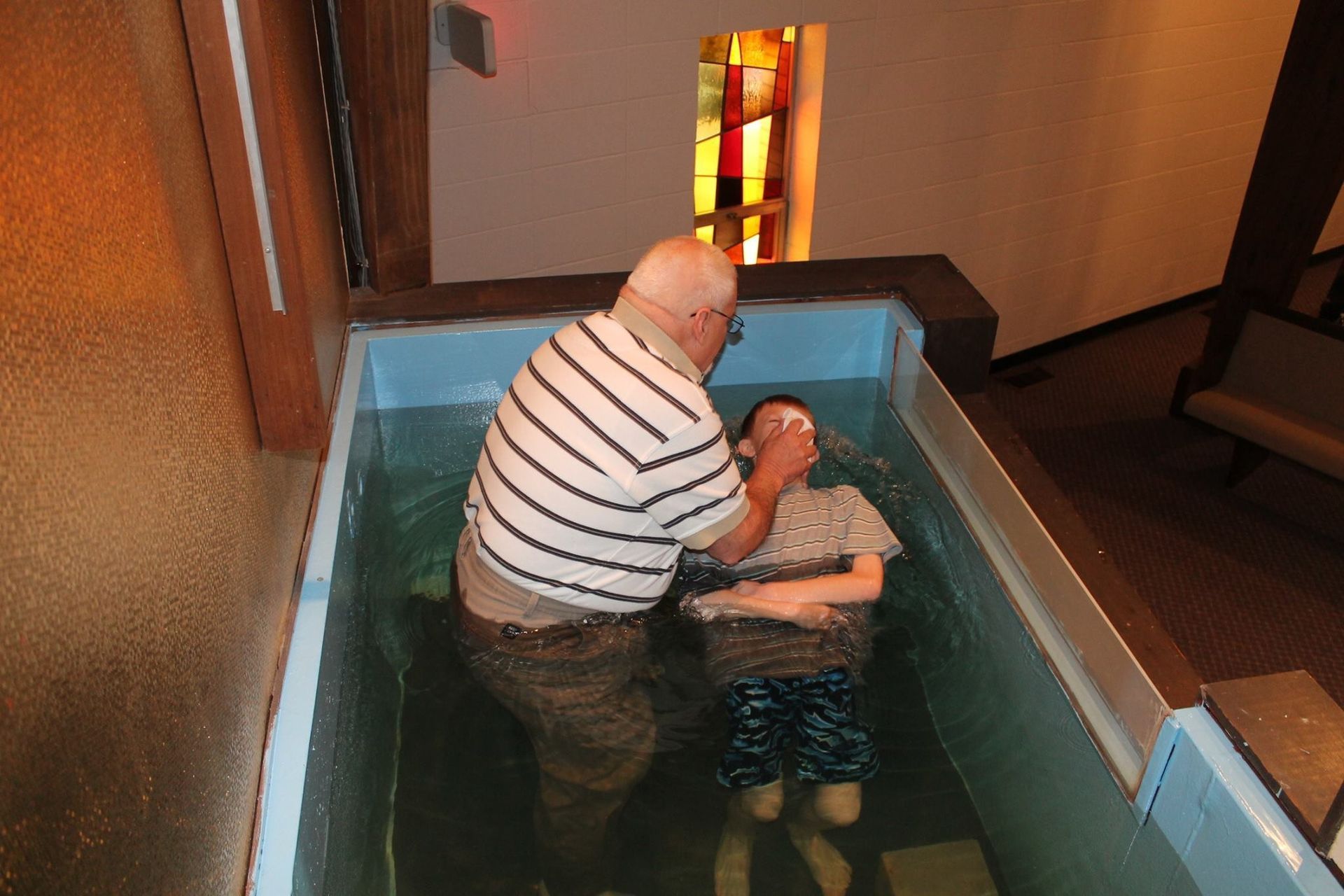 Person assisting another in a small indoor baptismal pool, with stained-glass light in the background