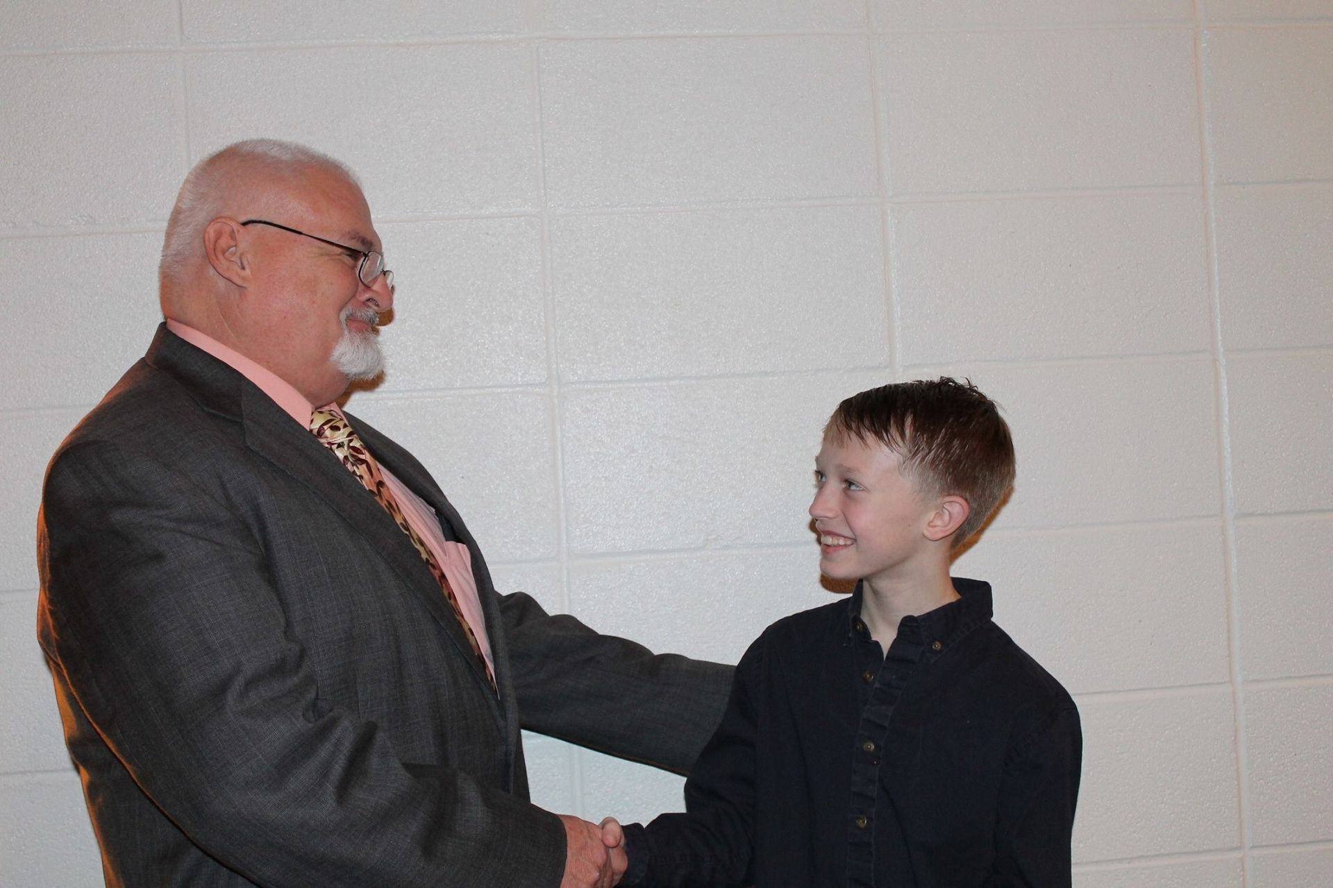 Two people in suits shaking hands and smiling in a hallway