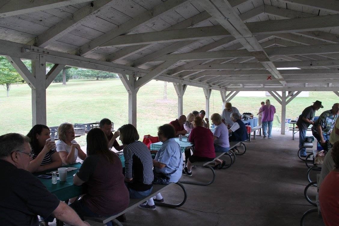 People seated at picnic tables under a wooden pavilion, gathered outdoors on a cloudy day.