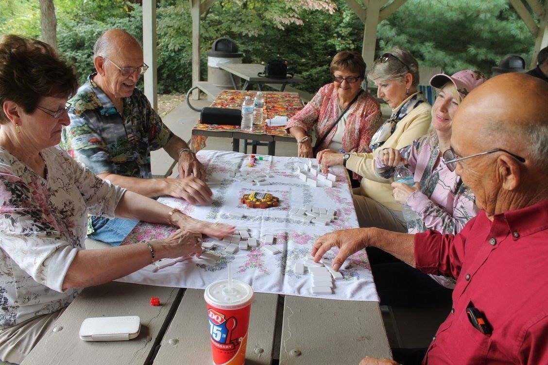 People seated at a picnic table under a pavilion, playing cards and talking with drinks on the table.