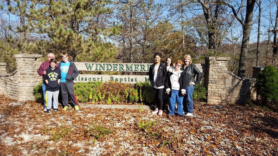 Group posing by a Windermere sign in a sunny wooded area with fallen autumn leaves.