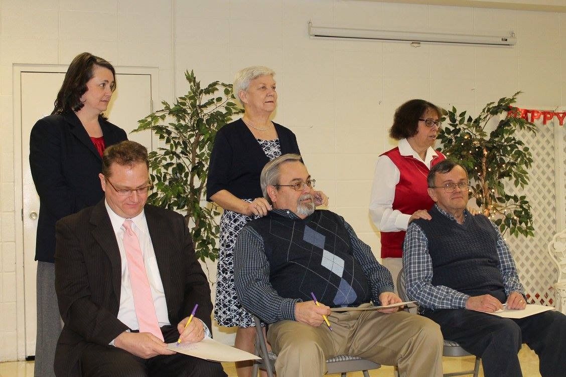 Six people seated indoors at an event, with two standing behind them near a plant and wall decor.