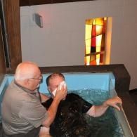 Man assisting a person in a baptismal pool inside a church, with stained glass in the background
