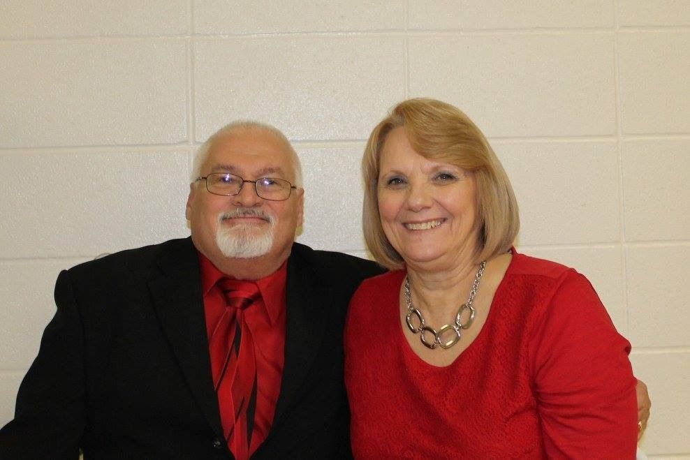Two people smiling side by side, wearing red and black, against a light wall.